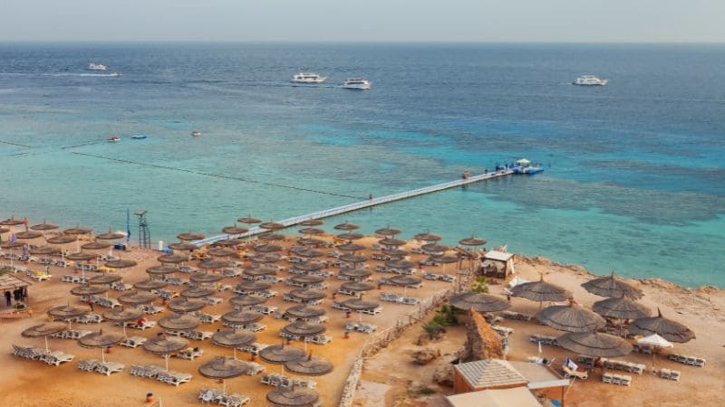 Beachfront lined with thatched umbrellas and sunbeds overlooking a turquoise sea with boats.
