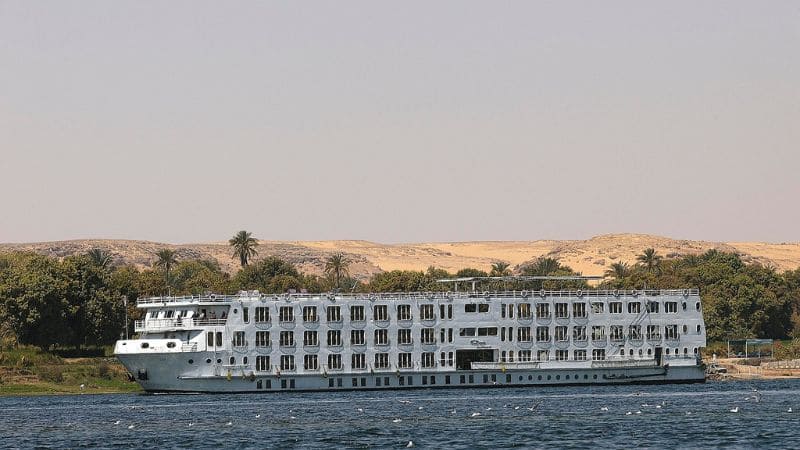 A white cruise ship sailing on the Nile River Cruises with desert hills in the background.