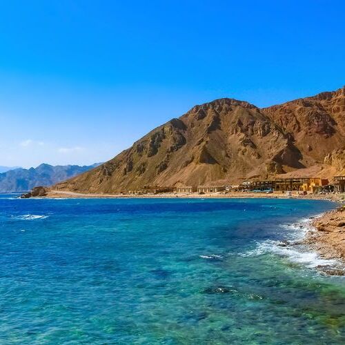 Coastal landscape with clear blue water, mountains in the background, and a strip of beachfront buildings under a bright blue sky