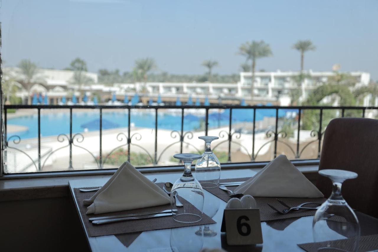An inviting dining table set for two with a clear view of a serene swimming pool and resort backdrop, captured from an indoor perspective.