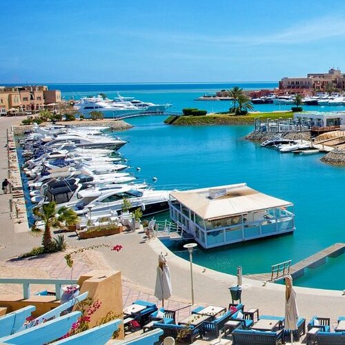 A panoramic view of a serene marina with clear blue waters, docked yachts, and surrounding colorful buildings under a bright blue sky