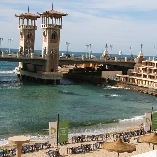 A tranquil beach setting with a distinctive dual-towered gatehouse leading to a pier, under clear skies.