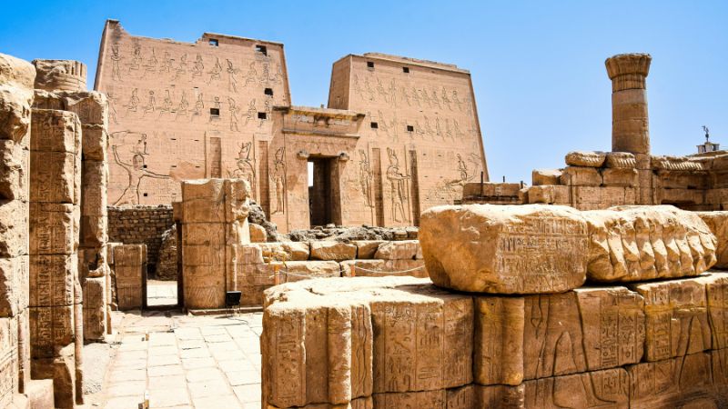 Ruins of an ancient Egyptian temple with large sandstone walls engraved with hieroglyphics and towering columns under a blue sky