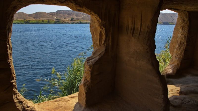 View through weathered stone windows overlooking a tranquil river and lush vegetation with distant hills under a clear sky
