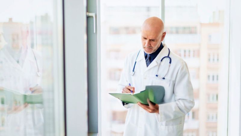 A male doctor in a white coat reviewing patient notes near a window with an urban backdrop