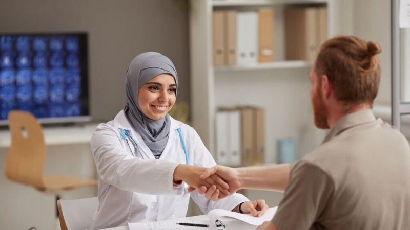 A smiling female doctor in a hijab shaking hands with a patient in a modern medical office