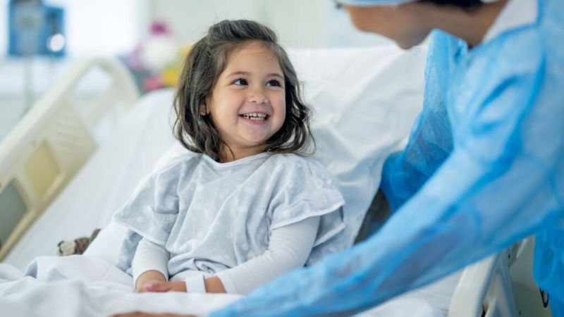 A young girl in a hospital bed smiling at a nurse in blue scrubs, creating a comforting and cheerful atmosphere