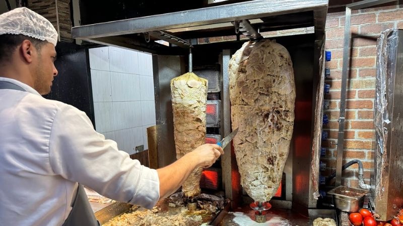 A chef slicing freshly cooked shawarma from a vertical rotisserie in a busy kitchen