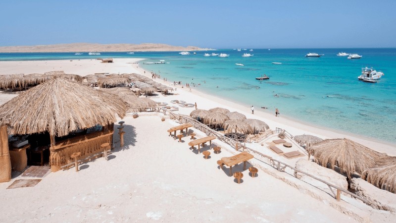 "Thatched huts line the sandy beach of a tropical island with clear turquoise waters and boats anchored nearby."