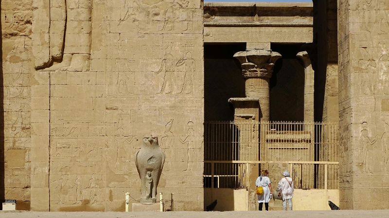 Entrance to an ancient temple with detailed carvings on the walls and two visitors in traditional hats observing the site