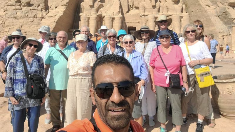 A group of tourists on a camel ride in front of the Great Pyramids of Giza, Egypt, with clear skies and desert sands in the background