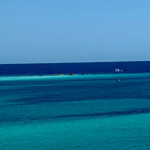 Graduated hues of blue in the sea with a distant sandy islet and a solitary boat under a clear sky
