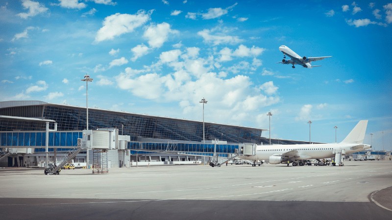 "Aircraft taking off near a modern airport terminal under a clear blue sky, with another airplane parked at the gate."