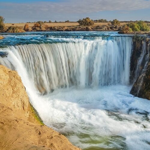 A wide waterfall with a heavy flow, surrounded by a semi-arid landscape under a clear sky.