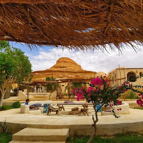 A tranquil courtyard with lush greenery, vibrant pink flowers, and a distinctive rock formation in the background, under a thatched roof