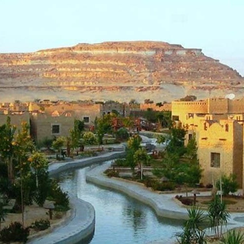 A tranquil water canal running through the Siwa Oasis with traditional mud-brick buildings and a grand mesa in the background.