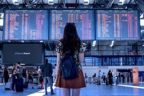 A woman standing in an airport, facing a large flight information display.