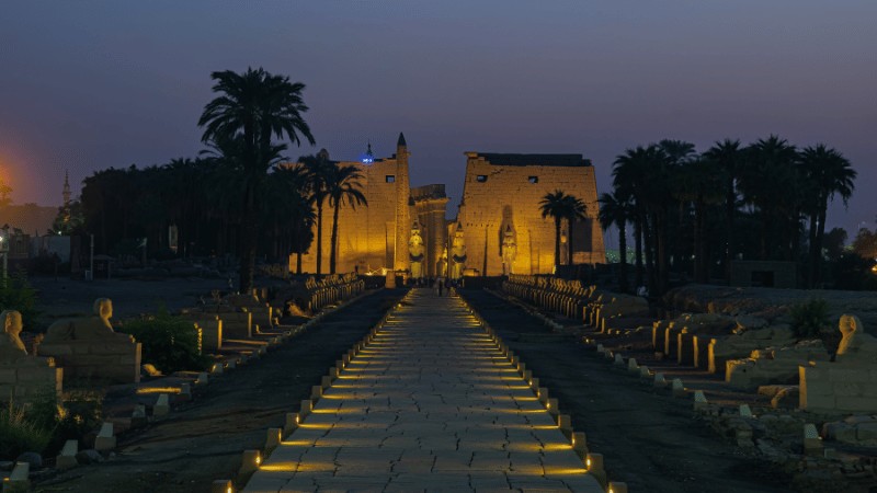 The Karnak Temple illuminated at dusk with a pathway lined by sphinx statues leading to the entrance