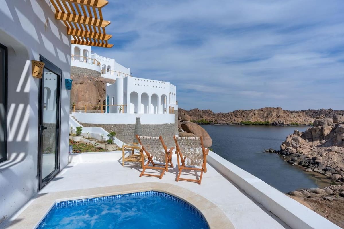 A white terrace with two wooden chairs overlooking a tranquil lake surrounded by rocky hills