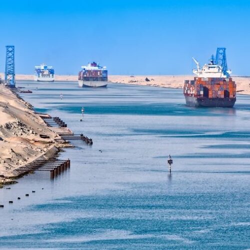 Cargo ships transit the narrow Suez Canal under a clear blue sky.