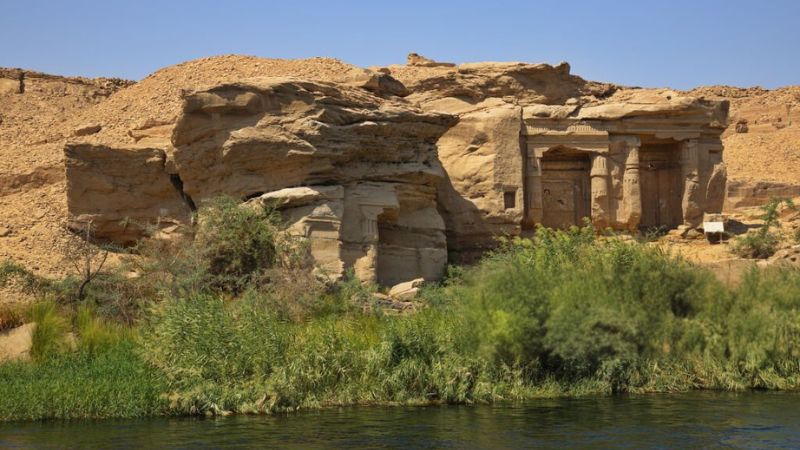 Small ancient temple ruins nestled into a rocky desert hillside, surrounded by greenery and overlooking a river