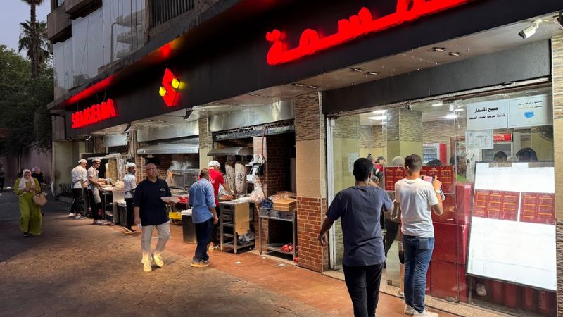Customers walking in and out of a busy shawarma shop with chefs preparing food outside