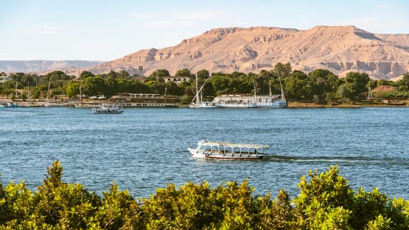 "A motorboat cruises along the Nile in Aswan, with lush greenery in the foreground and desert hills in the background