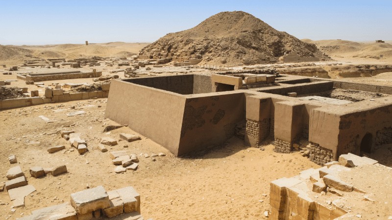"Sunlit ruins of ancient structures in a desert with sandy hills in the background."