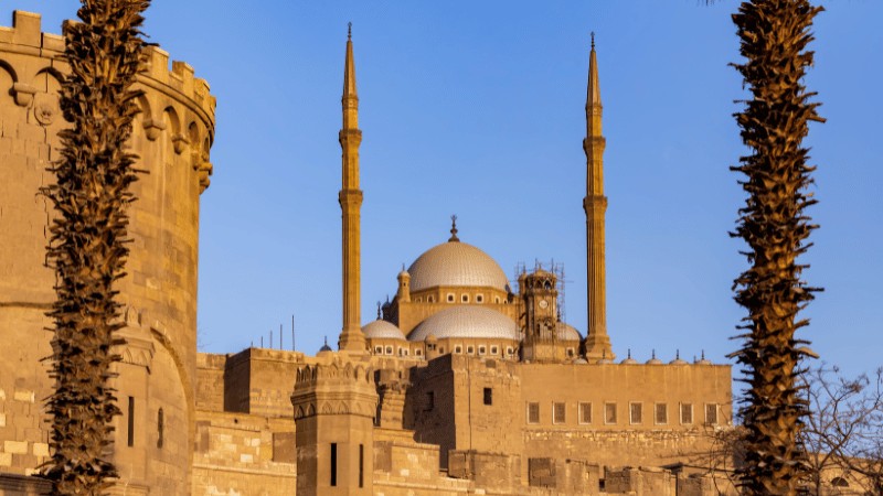 "Golden sunlight casting a warm glow on the Citadel of Saladin with surrounding palm trees in silhouette."