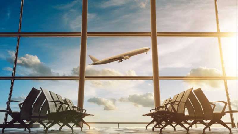 "An airplane taking off in the golden light of sunset, viewed from inside an airport terminal with empty seating in the foreground."