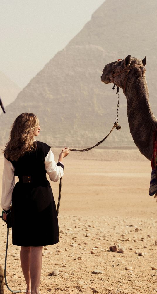 Woman standing with a camel in the desert with the Pyramids of Giza visible in the background